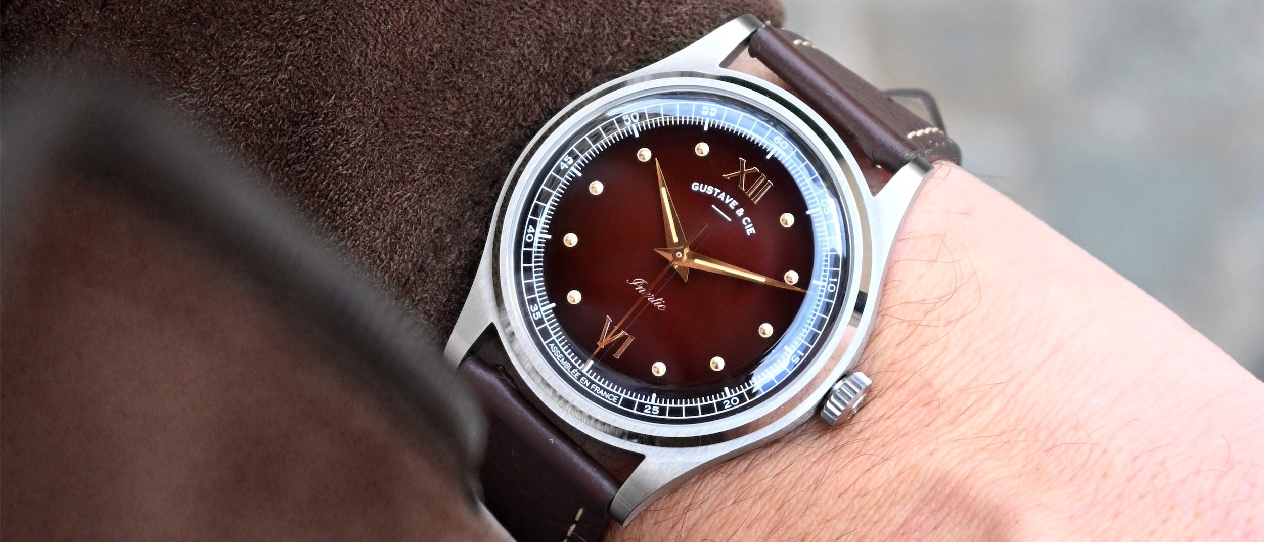 Montre automatique Gustave & Cie Léon 12H avec boîtier argent, cadran marron et bracelet cuir marron chocolat grainé, portée au poignet avec une veste en velours fine marron.