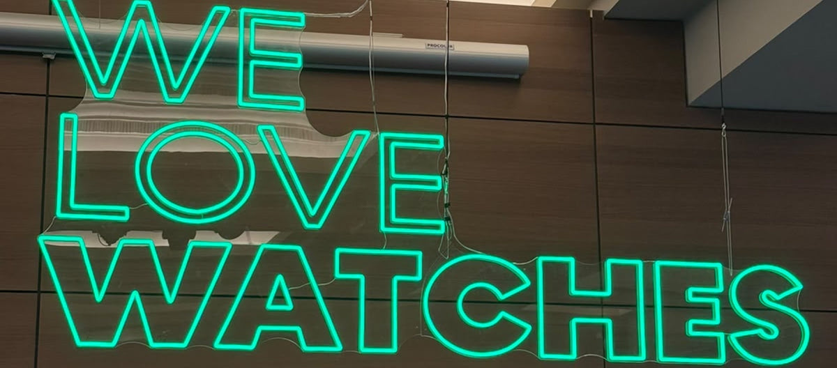 Enseigne lumineuse en néon vert affichant le texte 'WE LOVE WATCHES' accrochée sur un mur en bois foncé lors du salon.
