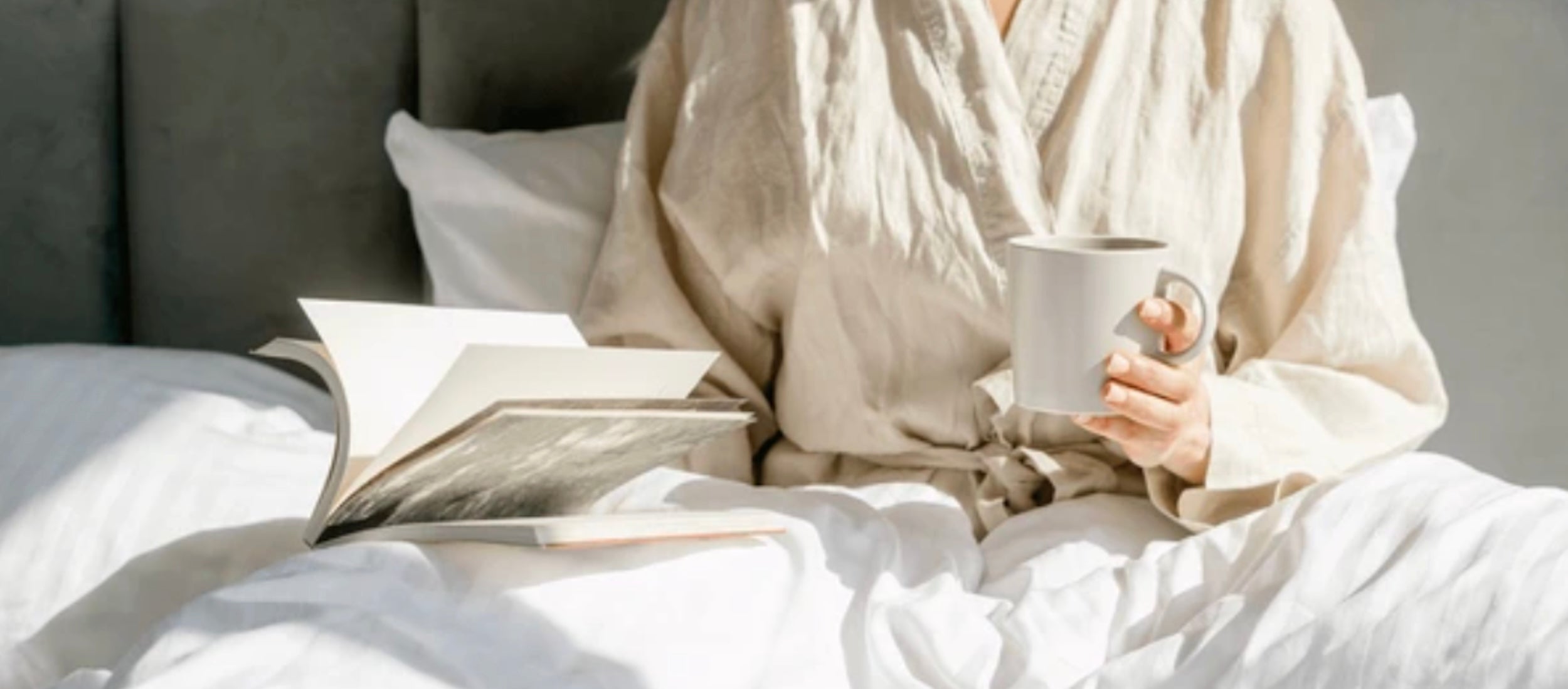 Une personne en peignoir, assise confortablement dans son lit avec un livre ouvert et une tasse, profitant d'un moment de détente matinale.