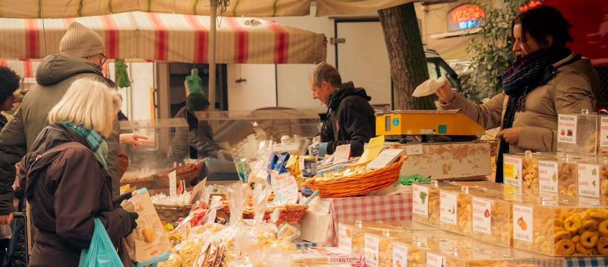 Commerçante pesant des produits sur une balance et clients achetant des gourmandises sur les étals d'un marché en plein air.