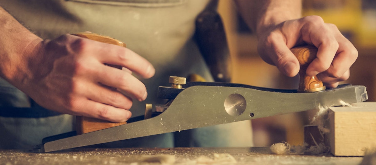 Mains d'un artisan utilisant un rabot manuel pour façonner une pièce de bois, soulignant la précision du geste et le travail du matériau.